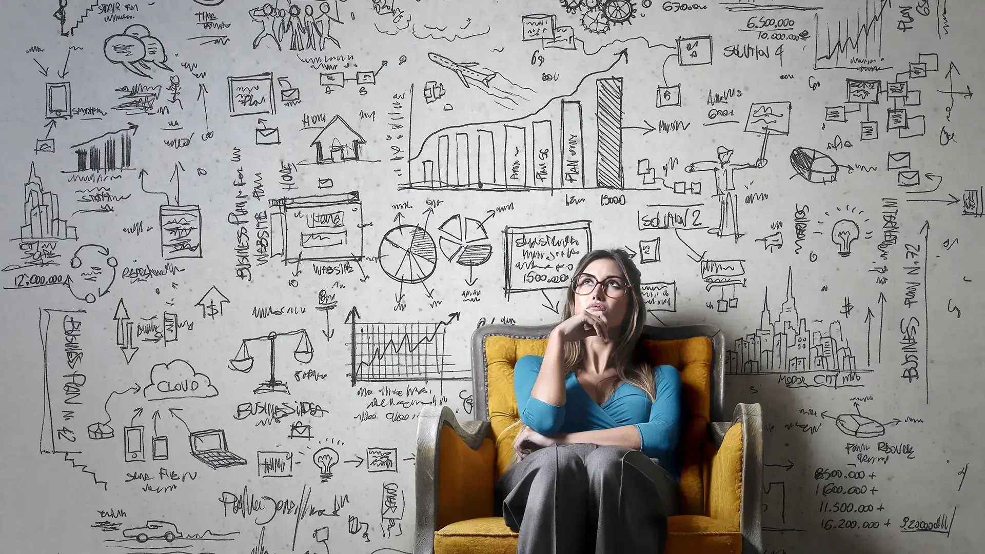 A woman in glasses, in a yellow chair, looking thoughtful with a background wall with concepts scribbled on it