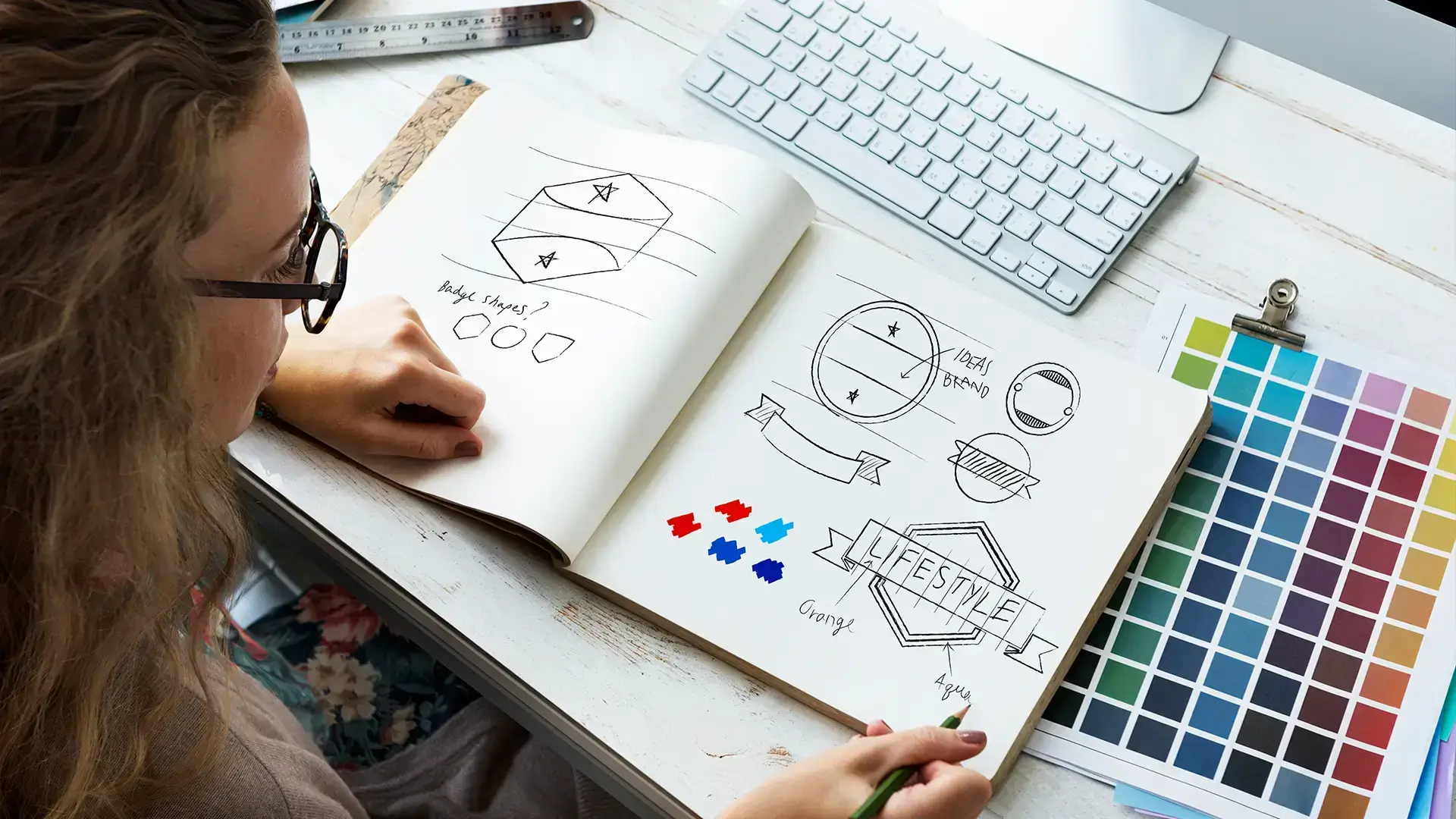 A woman at a desk scribbling in a notebook with a colour chart to her right