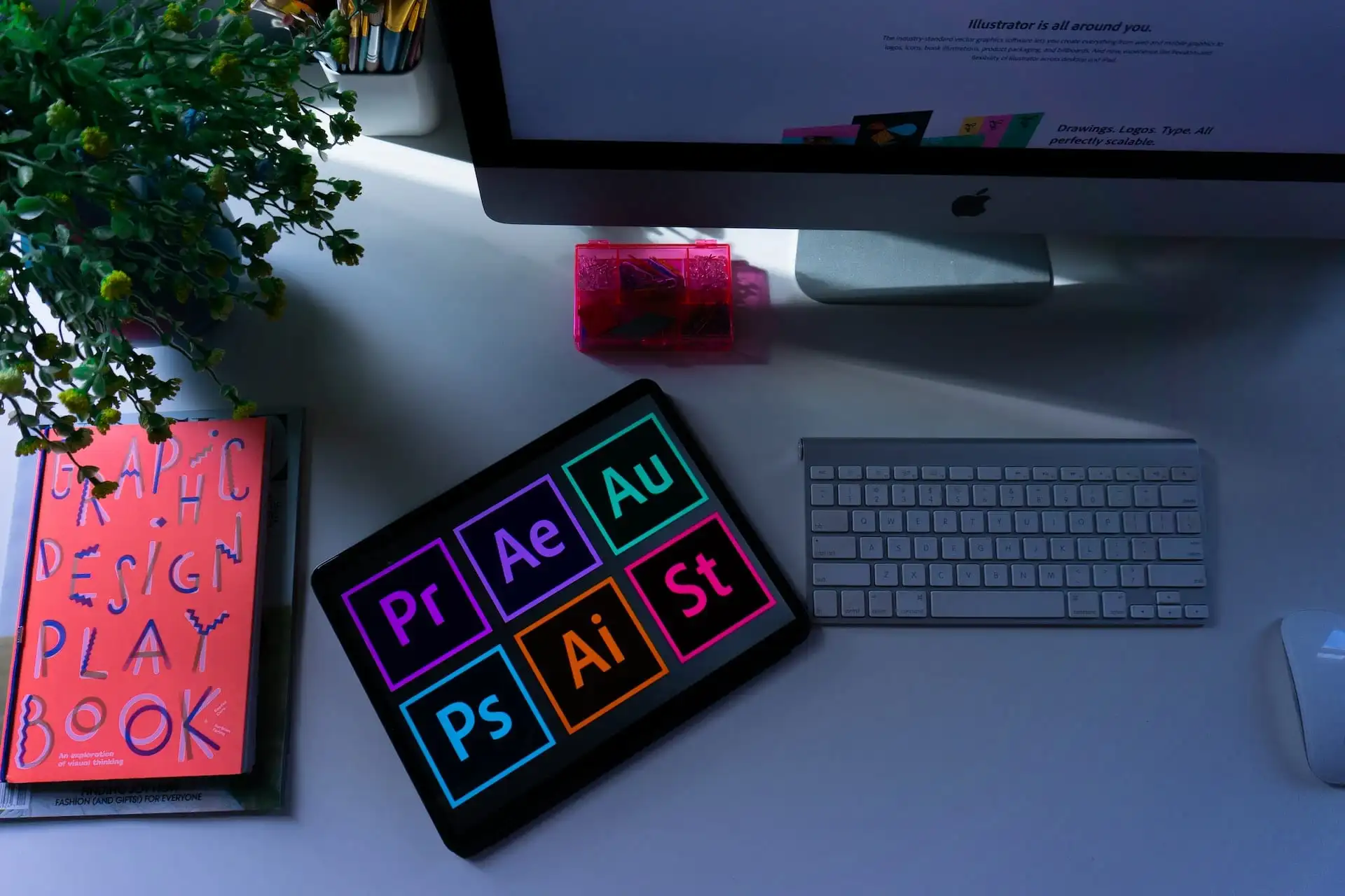 A mac screen and keyboard with to the side a notebook and screen showing the Adobe suite symbols